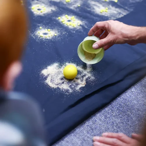 Kind streut mit einem Schälchen feinen Sand auf ein Sensorik-Tablett und experimentiert mit Mustern.