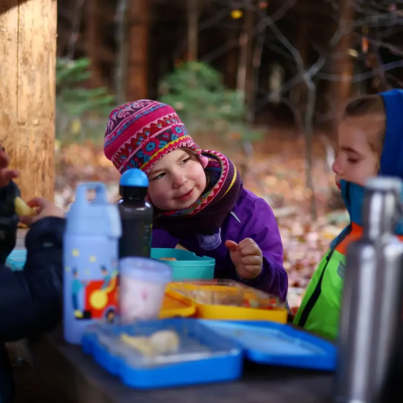 Kinder sitzen am Waldtisch und frühstücken mit Brotdosen und warmem Tee.