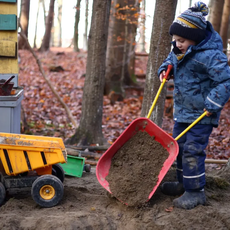 Kind schaufelt Erde mit einer großen Schaufel und spielt mit einem Bau-LKW.