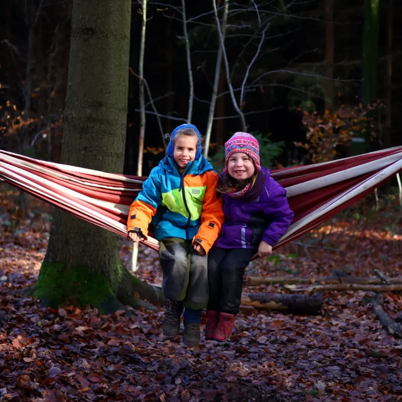 Zwei Kinder sitzen gemeinsam in einer Hängematte im Wald und lachen.