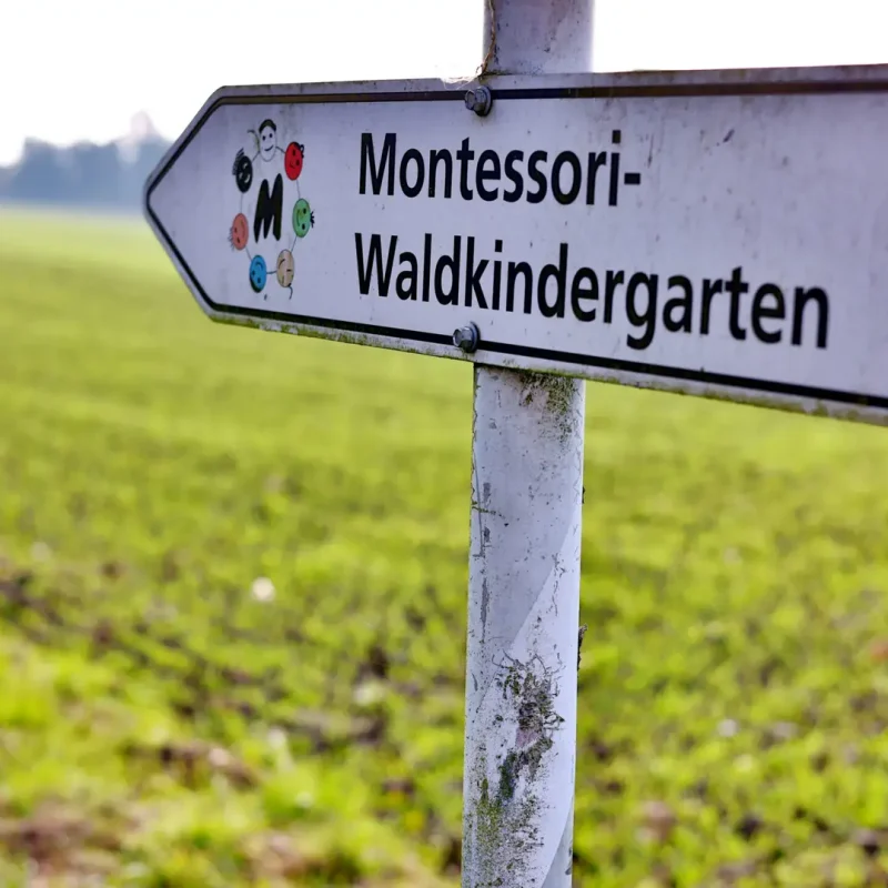 Wegweiser‐Schild zum Montessori-Waldkindergarten, aufgenommen am Rand einer grünen Wiese.
